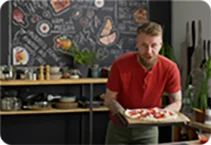 A man is holding a pizza with both of his hands in a kitchen environment.